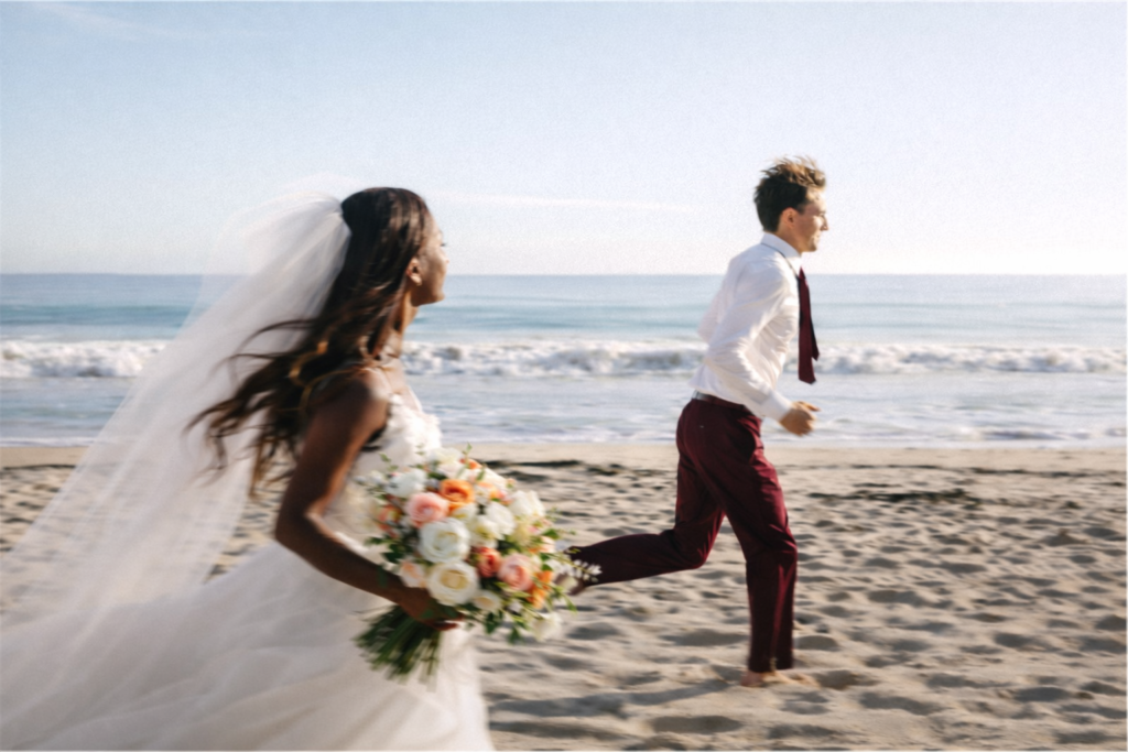 Bride holding a peach and white bouquet runs barefoot on the beach behind her groom after a seaside wedding ceremony.