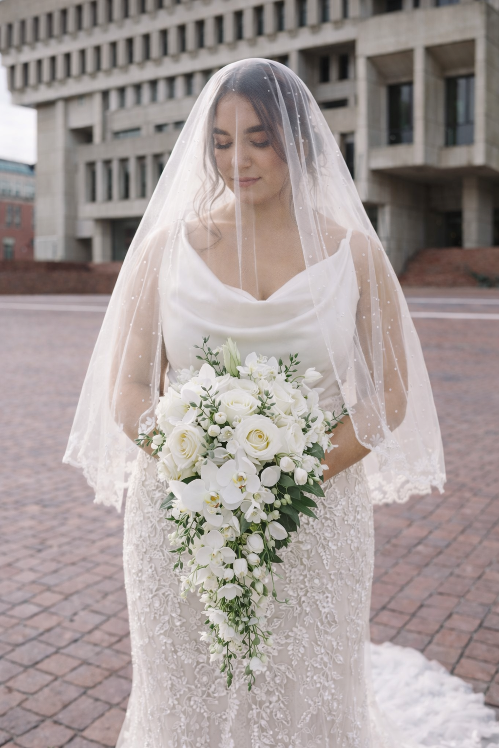A bride wearing a pearl-dotted face veil and satin cowl-neck top with a beaded lace skirt, holding a cascading white bouquet in a city setting.